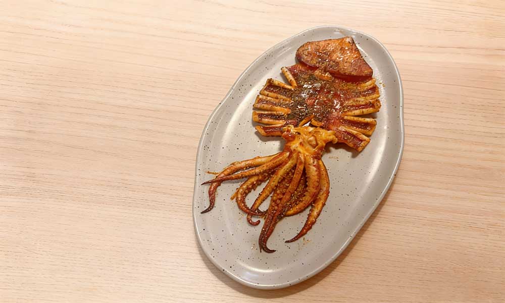 Whole grilled squid seasoned with cumin on speckled oval ceramic plate