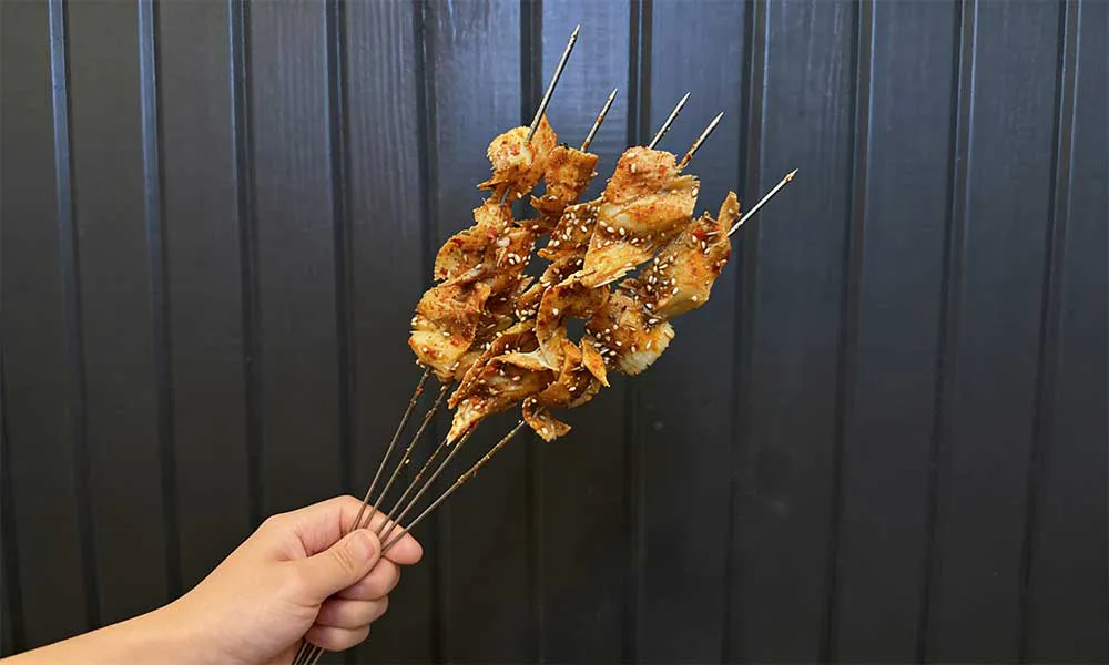 Hand holding spicy grilled tofu skin skewers sprinkled with sesame seeds against dark wall